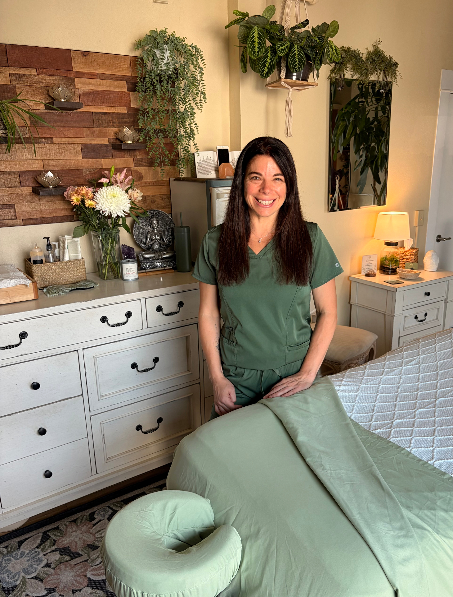 Cyndi Parris, LMT — standing beside the massage table in her plant-filled studio